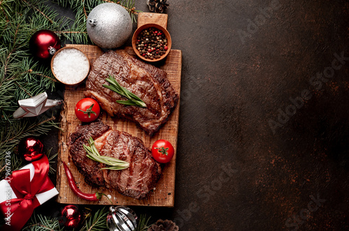 Christmas dinner for two, grilled beef steak, ribeye, greens and spices on a stone table with a Christmas tree and New Year's toys with copy space	