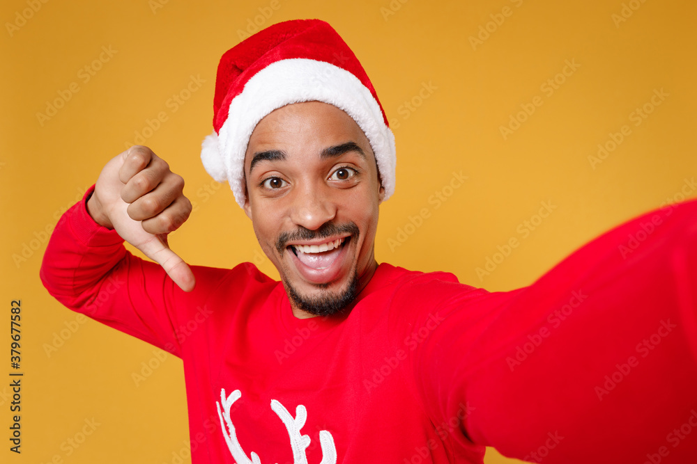 Close up funny young Santa african american man in Christmas hat doing selfie shot on mobile phone pointing thumb on himself isolated on yellow background. Happy New Year celebration holiday concept. Close up funny young Santa african american man in Christmas hat doing selfie shot on mobile phone pointing thumb on himself isolated on yellow background. Happy New Year celebration holiday concept.