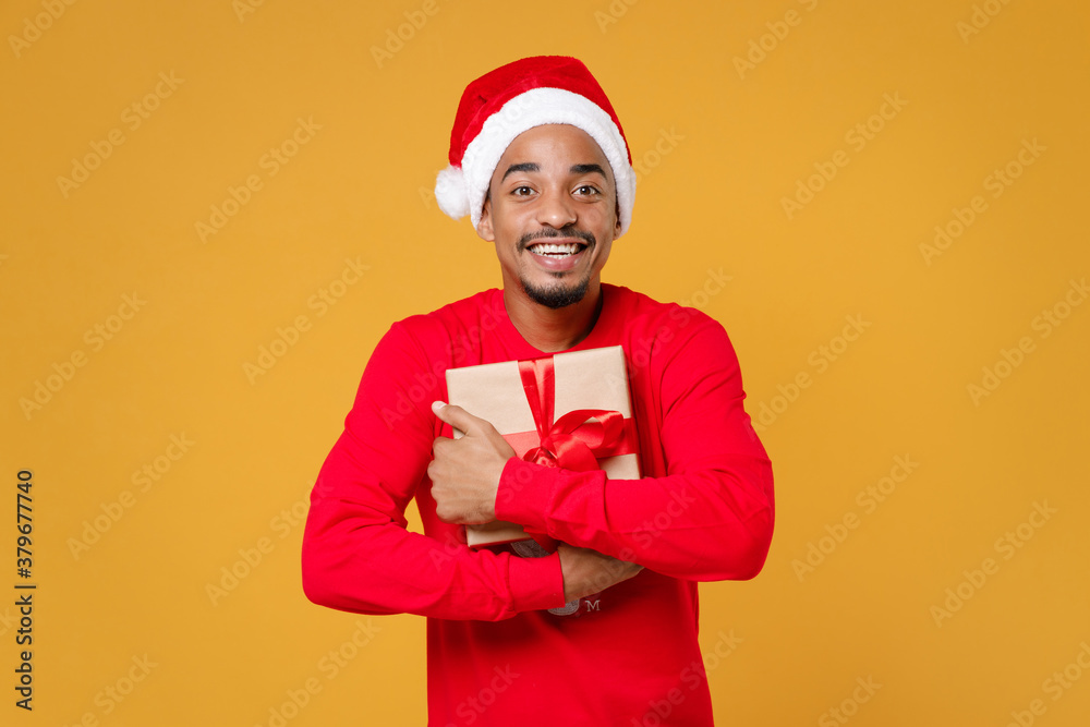 Cheerful Santa african american man in red sweater Christmas hat hold present box with gift ribbon bow isolated on yellow background studio portrait. Happy New Year celebration merry holiday concept. Cheerful Santa african american man in red sweater Christmas hat hold present box with gift ribbon bow isolated on yellow background studio portrait. Happy New Year celebration merry holiday concept.
