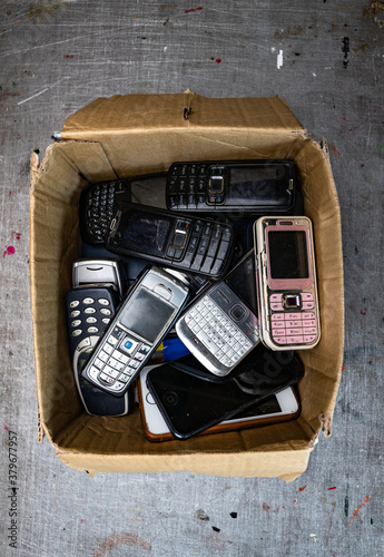 old, obsolete and broken cellphones in a cardboard box. scratched metal background.