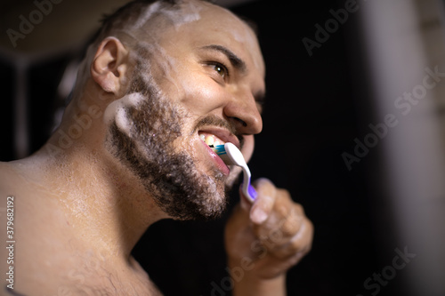 close up portrait of young bearded naked brunette man brushes his teeth, soaping his head and beard with foam of hair care product or antihairloss remedy taking shower on black backdrop photo