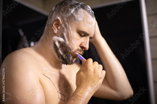 young bearded brunette man with tattoo on his naked chest washed his head and beard with shampoo or antihairloss remedy and brushed his teeth taking shower on black backdrop. man rushes to work photo