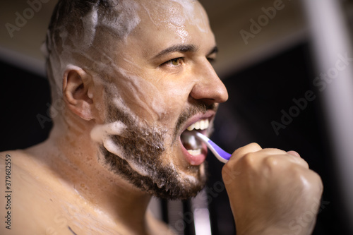 young bearded naked brunette man brushes his teeth, soaping his head and beard with hair care product taking shower. antihairloss remedy concept. man rushes to work and takes quick shower photo