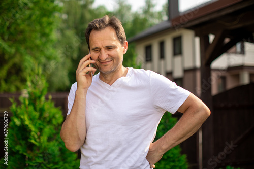 Happy middle aged man talking by smartphone, smiling friendly man standing outdoors on summer sunny day with mobile phone. Copy space, communication, digital people, mobile operator ads concept