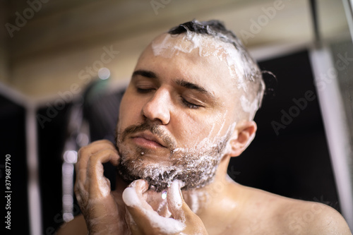 young bearded naked brunette man washes his beard and head with shampoo foam. Advertising for men's hair cosmetics or conditioner. beard care concept, antihairloss remedy. anti-hair loss application