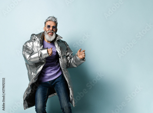 Mature man in purple pullover, sunglasses, silver colored down jacket, jeans. Running, posing on blue background