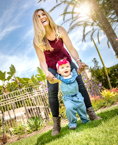 Adorable image of blonde mother helping her baby daughter to walk at a park with palm trees on a sunny day with blue sky and white clouds photo