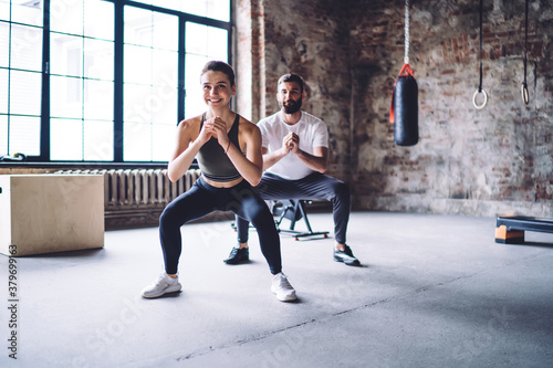Happy slim male and female couple in love squatting together keeping perfect body shape, smiling woman and man training muscles and flexibility losing weight on training in modern designed gym