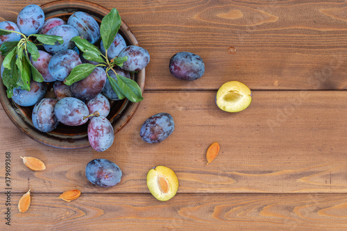 A few washed ripe blue plums with leaves in a brown plate on the wooden table. Several seeds and halves of fruit lie separately. The view from the top