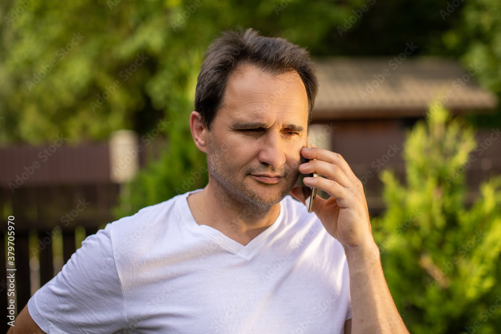 Calm portrait of positive unshaven mature European 40s businessman in white T-shirt speaking on mobile phone outdoor on blurred green natural background. Business concept for Controlling feelings Calm portrait of positive unshaven mature European 40s businessman in white T-shirt speaking on mobile phone outdoor on blurred green natural background. Business concept for Controlling feelings