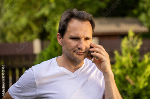 Calm portrait of positive unshaven mature European 40s businessman in white T-shirt speaking on mobile phone outdoor on blurred green natural background. Business concept for Controlling feelings