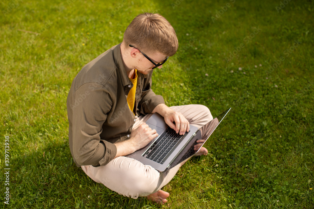 Young millennial man in casual wear sitting on grass in park working on laptop, checks email, coffee break, digital addiction, remote work, modern freelance lifestyle concept, top view, copy space Young millennial man in casual wear sitting on grass in park working on laptop, checks email, coffee break, digital addiction, remote work, modern freelance lifestyle concept, top view, copy space