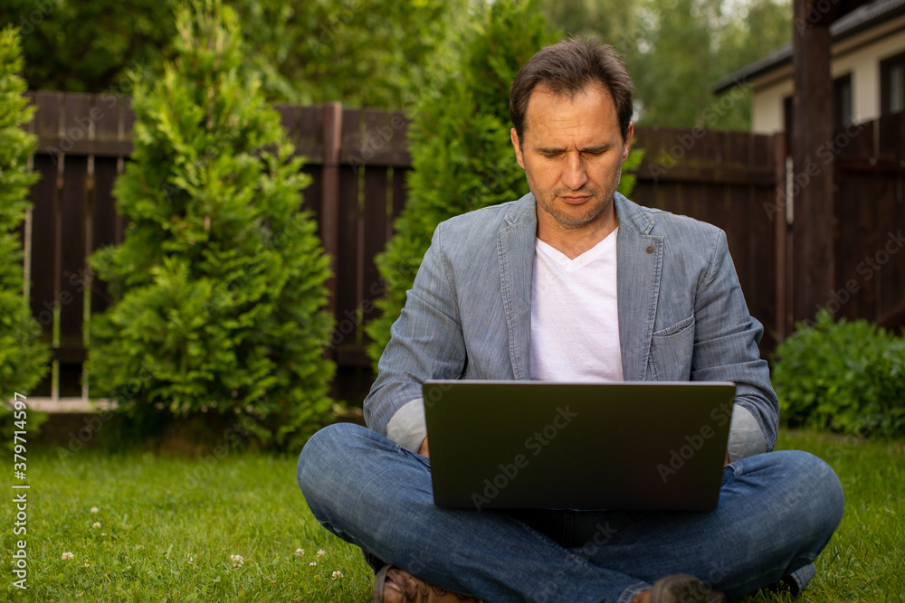 Focused unshaven mature European 40s businessman sits with crossed legs on grass with laptop on blurred green natural background. Distance work concept Focused unshaven mature European 40s businessman sits with crossed legs on grass with laptop on blurred green natural background. Distance work concept