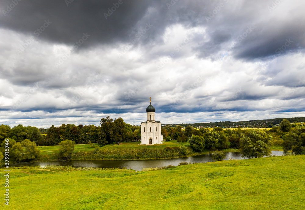 a panoramic view of the white old church on a green meadow between the tributaries of the river against the backdrop of thunderclouds filmed from a drone