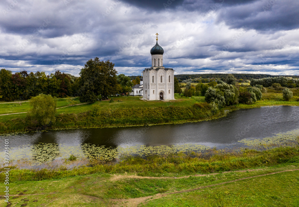 a panoramic view of the white old church on a green meadow between the tributaries of the river against the backdrop of thunderclouds filmed from a drone