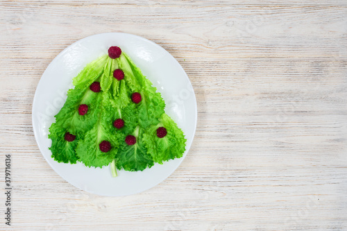 The Christmas tree is lined with green lettuce leaves, decorated with red raspberries on a white plate. Food for the New Year. Table decoration. View from above. Place for an inscription.
