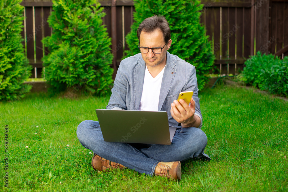 Serious adult man sitting at lawn outdoors, attractive male freelancer working on computer laptop and typing on mobile phone, businessman working from home on summer morning. Remote work, lifestyle