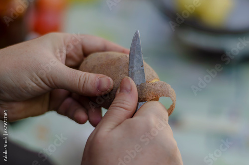 An adult woman peels raw potatoes with a kitchen knife.