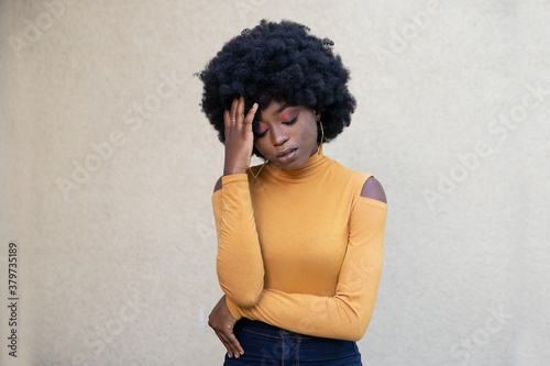 Horizontal shot of frustrated African American woman with curly hair and closed eyes, suffers from headache, experiencing stress and tension, wearing casual wear, posing outdoors on light background. © Larysa