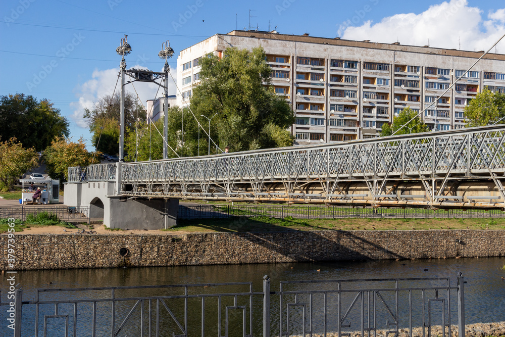 View on pedestrian bridge in Ivanovo with metal posts, fence  and light lamps under blue sky, river, city buildings