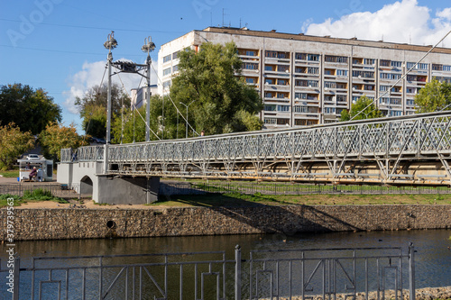 View on pedestrian bridge in Ivanovo with metal posts, fence and light lamps under blue sky, river, city buildings