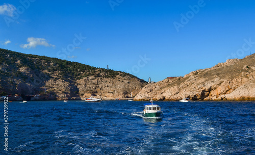 white yacht sails in blue sea among big red stones cliffs, mountains, summer light of sun, vacation