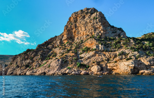 red orange stone rock cliff stands on shore of the blue sea on a bright sunny day, summer sky with clouds