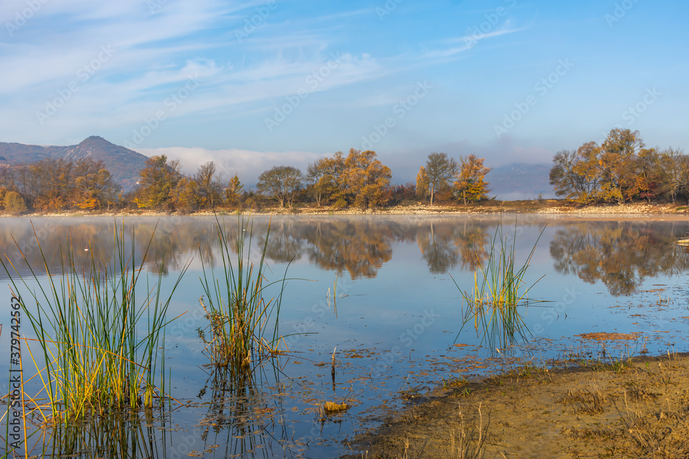 Fototapeta premium Beautiful autumn landscape with yellow trees and their reflection in the lake. Bright Sunny autumn day. Yellow trees in a blue sky. Horizontal panorama with a light haze. A little fog over the water.