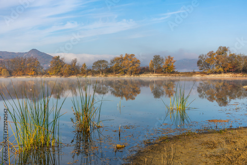 Beautiful autumn landscape with yellow trees and their reflection in the lake. Bright Sunny autumn day. Yellow trees in a blue sky. Horizontal panorama with a light haze. A little fog over the water.