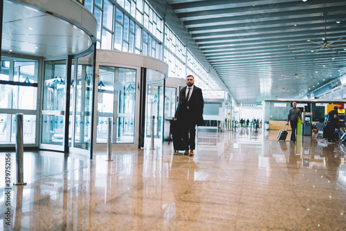 Full length portrait of formally dressed corporate director with luggage suitcase walking inside of airport and looking at camera, proud CEO posing during business trip searching departure gate © BullRun