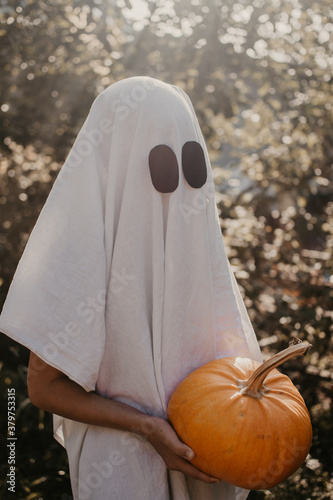 Halloween ghost costume in vintage style. Suit made of sheets. Child in a party costume scares. Trick or treat. Young child dressed as a ghost holding a pumpkin for halloween.