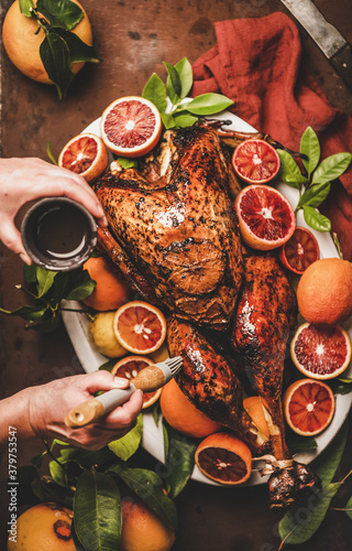 Christmas or Thanksgiving Day festive table preparation. Womans hand putting olive oil on whole roasted turkey with citrus fruit over rusty table background, top view. Holiday gathering food concept