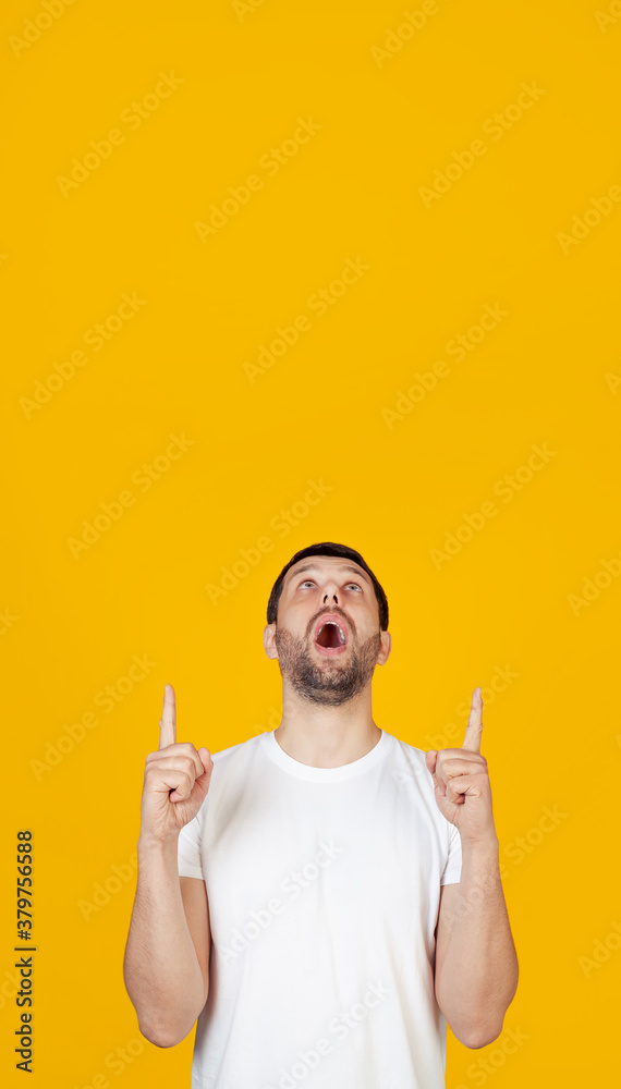A young man with a beard in a white T-shirt is amazed and surprised, looking up and pointing with his fingers and raised hands. Stands on isolated yellow background A young man with a beard in a white T-shirt is amazed and surprised, looking up and pointing with his fingers and raised hands. Stands on isolated yellow background