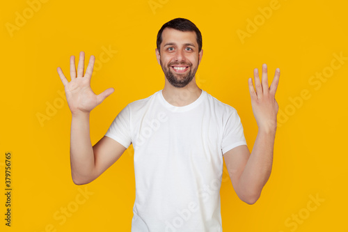 Modern young man with a beard in a white tank top shows number nine with fingers on hand smiling confidently and happily looking into the camera. The man shows nine fingers. Number 9 yellow background