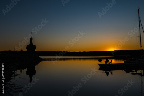 The silhouette of a church, a fisherman and a boat against the backdrop of the sunset. Night, reflection in the water. Dark silhouettes of the chapel.