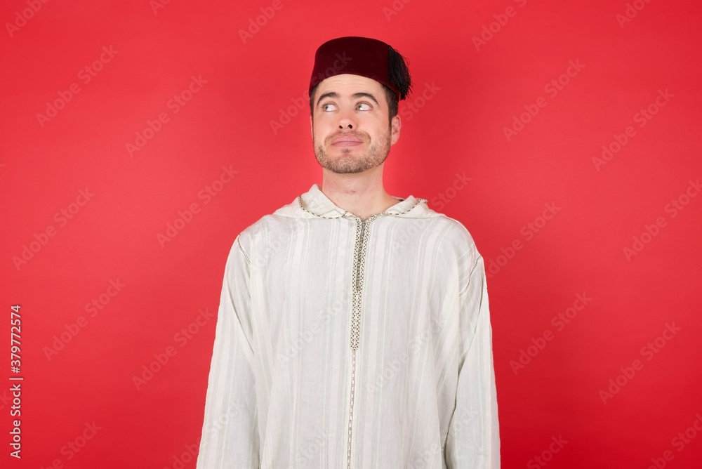 Photo of cheerful positive cute young caucasian muslim man wearing djellaba and traditional hat over red background, looking aside into empty space thoughtful Photo of cheerful positive cute young caucasian muslim man wearing djellaba and traditional hat over red background, looking aside into empty space thoughtful