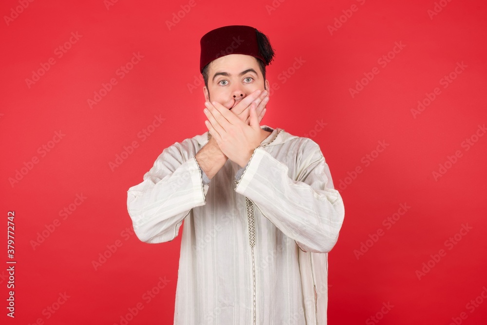 young caucasian muslim man wearing djellaba and traditional hat over red background shocked covering mouth with hands for mistake. Secret concept. young caucasian muslim man wearing djellaba and traditional hat over red background shocked covering mouth with hands for mistake. Secret concept.