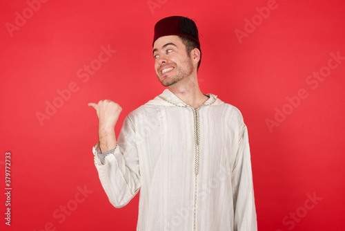 young caucasian muslim man wearing djellaba and traditional hat over red background promoter looking at copy space having advertisements  photo