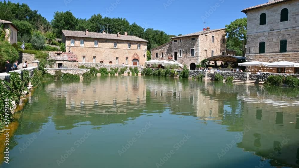 Bagno Vignoni, Tuscany, Italy. August 2020. View on the water mirror of the spring basin: here thermal water flows which fills the rectangular basin. People walk the perimeter. Nice sunny day. Bagno Vignoni, Tuscany, Italy. August 2020. View on the water mirror of the spring basin: here thermal water flows which fills the rectangular basin. People walk the perimeter. Nice sunny day.