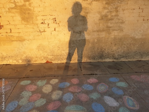 The shadow of a woman against a wall on a footpath covered with children's chalk drawings. 