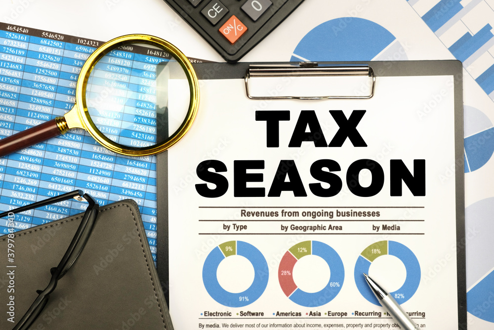 The businessman has on his desk graphs with reports, a notebook, a magnifying glass and a document with the inscription - TAX SEASON
