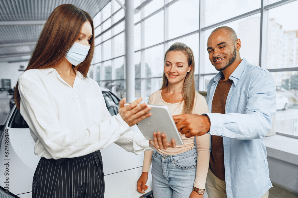 Fototapeta premium Car saleswoman wearing medical mask shows buyers couple something on digital tablet