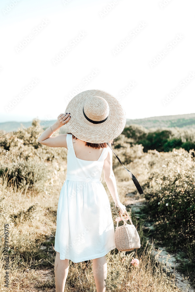 a girl on a walk goes among the trees. the view from the back, dressed in a white dress. a hat on his head, flowers in his hands
