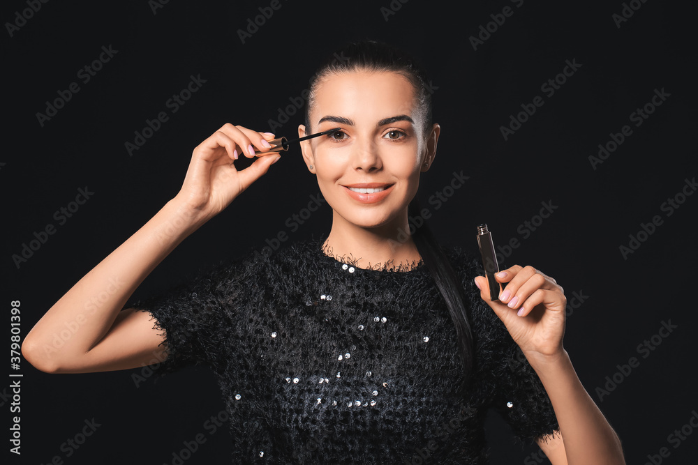 Beautiful young woman applying mascara against dark background Beautiful young woman applying mascara against dark background