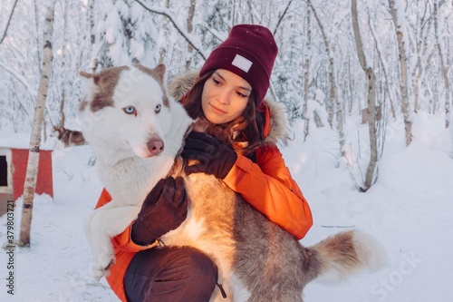 girl in a bright orange jacket sits and plays and hugs with a husky breed dog in the winter forest after snowfall during frost in the rays of the bright sunset sun