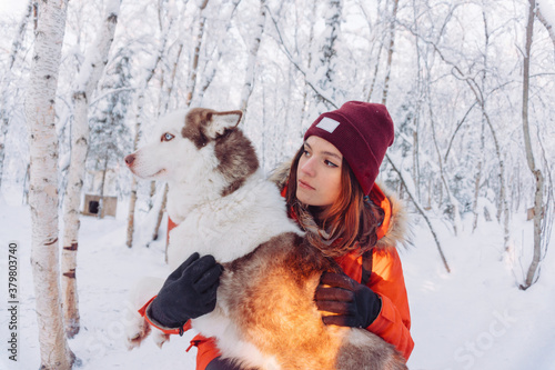 girl in a bright orange jacket sits and plays and hugs with a husky breed dog in the winter forest after snowfall during frost in the rays of the bright sunset sun