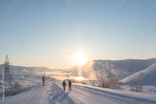 two girls in bright jackets walk along a white snowy road in the rays of the bright orange sun through the tundra beyond the Arctic Circle on a frosty clear day