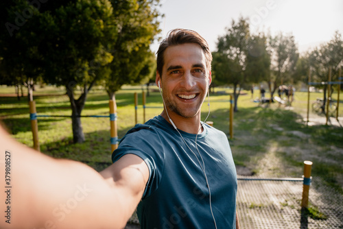 Smiling young male athlete takes photo of self before exercising outdoors in the sunny park listening to music in ear phones photo