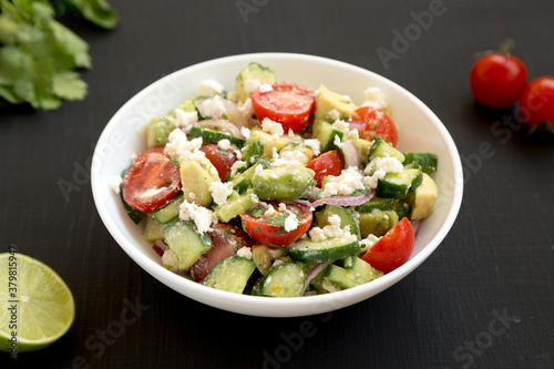 Delicious Avocado Tomato and Cucumber Salad in a white bowl on a black background, side view. Close-up.