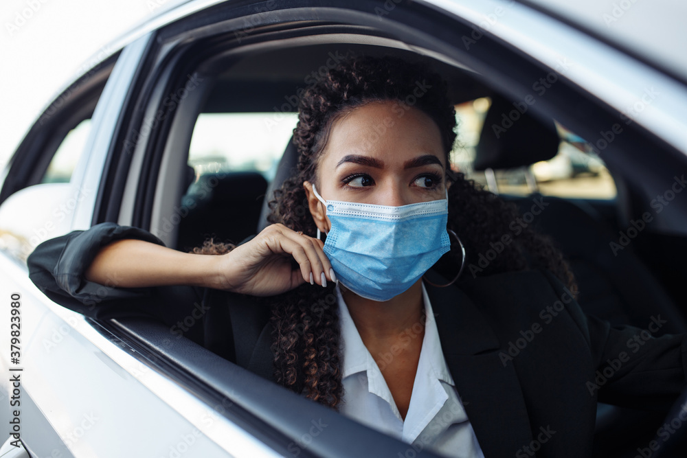 Young business woman fixing and adjusting her medical mask while sitiing in the car behind the steering wheel. Business trips during pandemic, new normal and coronavirus travel safety concept. Young business woman fixing and adjusting her medical mask while sitiing in the car behind the steering wheel. Business trips during pandemic, new normal and coronavirus travel safety concept.